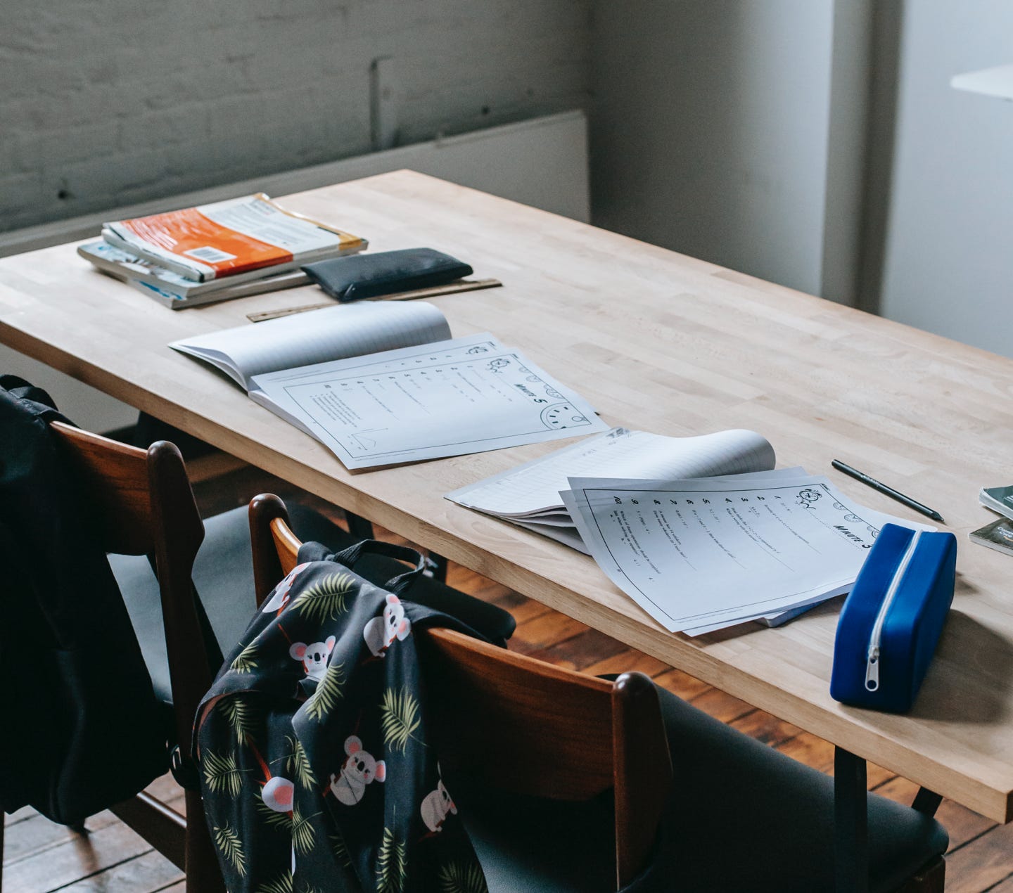 Wooden desk with opened copybooks and workbooks placed near chairs with backpacks in light classroom